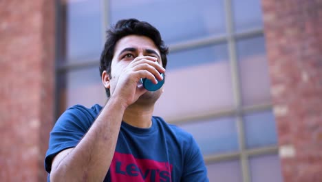 young south asian man drinks water from a bottle cap sitting outside during the day - low angle shot