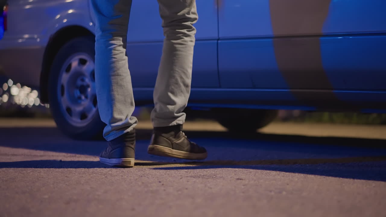 primer plano de una calle nocturna con pies caminando hacia un coche, mensajero adulto en vaqueros y zapatillas pisando asfalto irregular junto a un sedán plateado, luces de neón azules y faros proyectando reflejos en el panel, ángulo bajo
