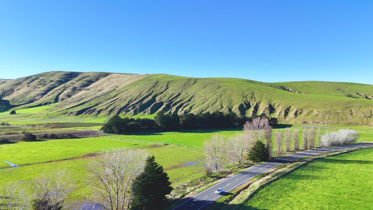 Aerial view of a winding road through lush green hills under clear blue skies in Akaroa, New Zealand