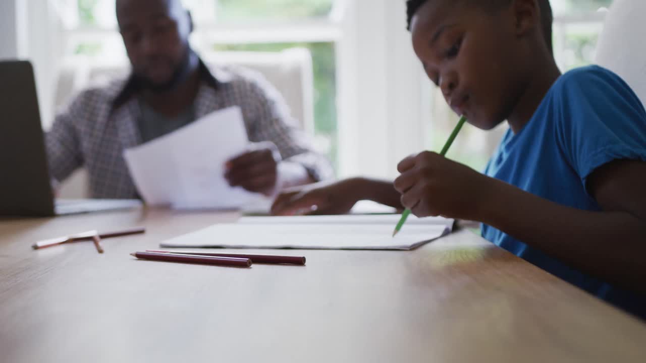 African american son sitting at kitchen table doing school work with father working in background