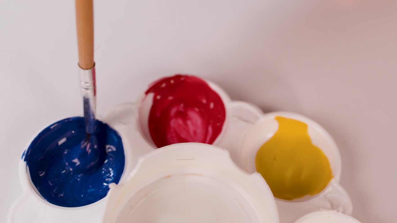 Hand uses brush to mix blue paint in palette, vibrant colors, overhead view, bright lighting