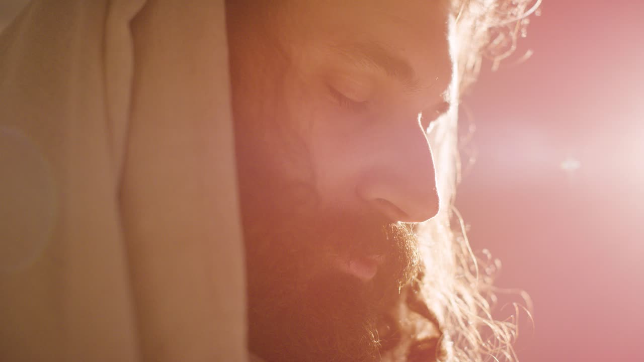 Backlit Close Up Of Man Wearing Robes With Long Hair And Beard Representing Figure Of Jesus Christ Praying