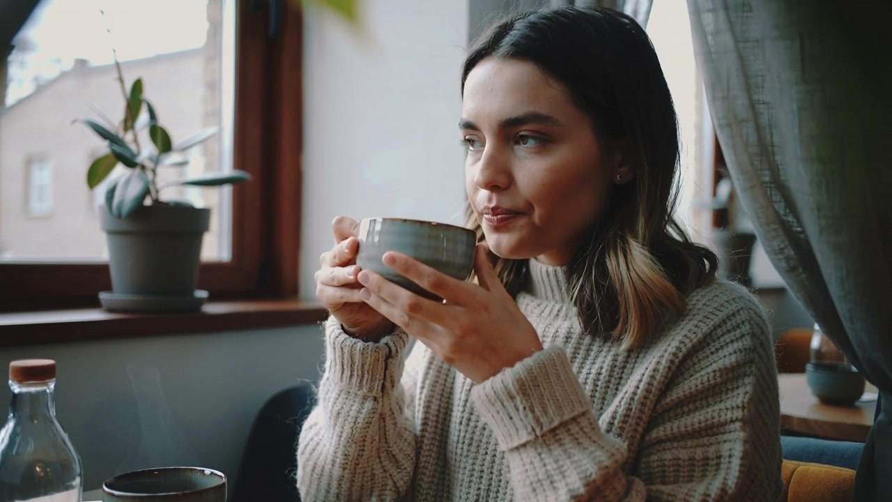 Deteniéndose a pensar, una mujer con suéter color crema levanta una gran taza de cerámica junto a la ventana, bebiendo a sorbos