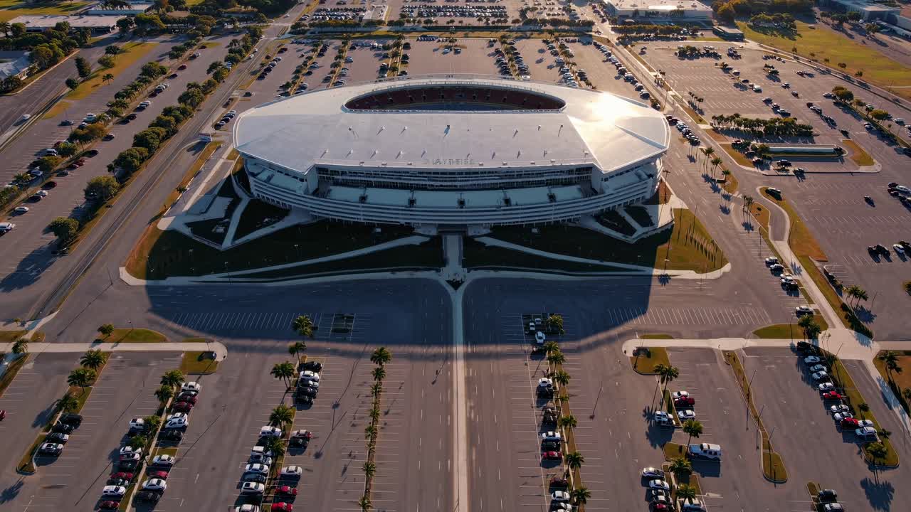 Aerial view of a modern stadium surrounded by expansive parking lots, showcasing the architectural design and vibrant atmosphere of a bustling event space with clear skies