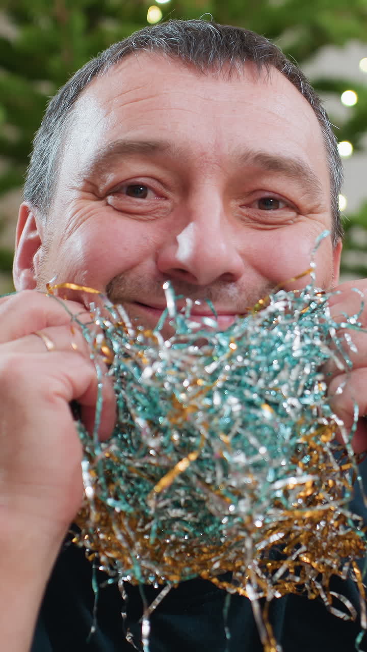 Крупный план: мужчина европеоидной расы держит синюю с золотом мишуру, задумчивый декоратор, которого затем что-то забавляет, распутывает гирлянду возле освещенной ели, мягкий эффект боке, блеск на щетине, кадры показывают старательного мастера за работой