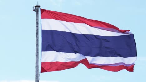 thailand national flag hoisted on a white tall steel pole blown by the wind creating an animated wave on a backdrop of a clear blue sky and white clouds