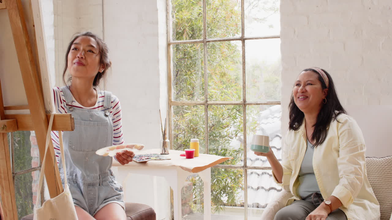 Painting on canvas, multiracial young woman while grandmother enjoys coffee, both smiling, at home