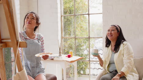painting on canvas, multiracial young woman while grandmother enjoys coffee, both smiling, at home