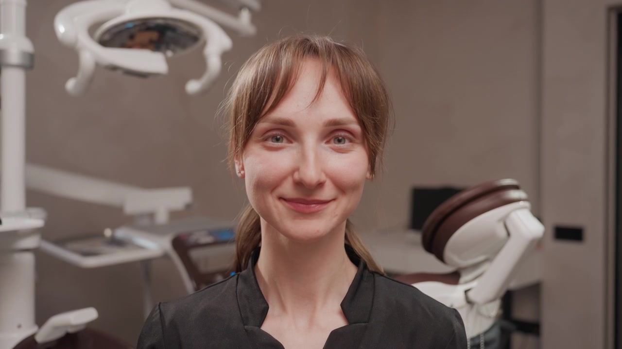 Närbild på tandhygienist som bär skyddande ansiktsskydd på klinik, justerar och tar bort genomskinligt visir medan hon står nära patientstol och taklampa. Tandläkarmiljö.