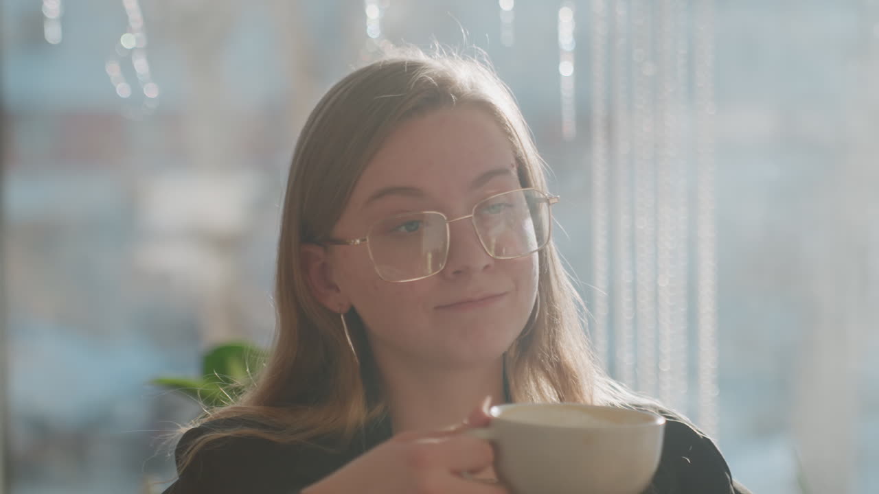 Jovem estudante caucasiana sorrindo com café perto da janela, pausa casual para estudo, luz solar suave através do vidro, plantas e interior acolhedor, expressão relaxada e riso suave, rotina confortável de café.