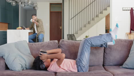 Using VR headset, boy lying on couch while Indian father working in background