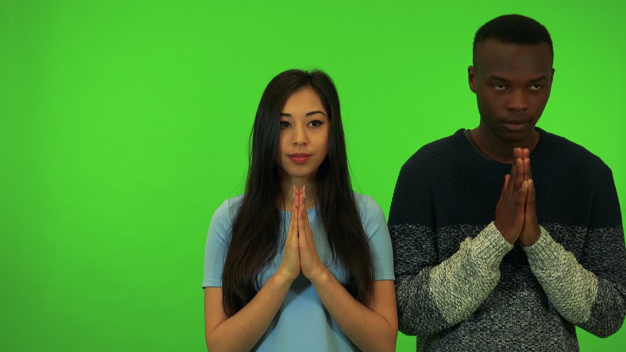 A young Asian woman and a young black man pray with hands clasped together - green screen studio
