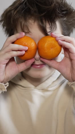 handsome teenage kid sitting in front of camera. kid puts some tangerines as his eyes. close up. vertical video