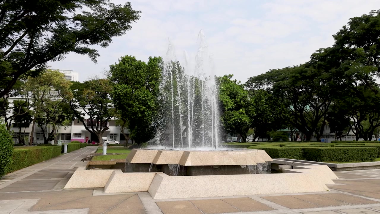 Una toma amplia captura una fuente de agua escalonada rociando hacia arriba en un tranquilo parque universitario.