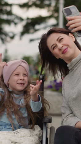 mamma takes selfies while little girl with disability making funny gestures with hands. woman enjoys spending time with daughter in wheelchair in park