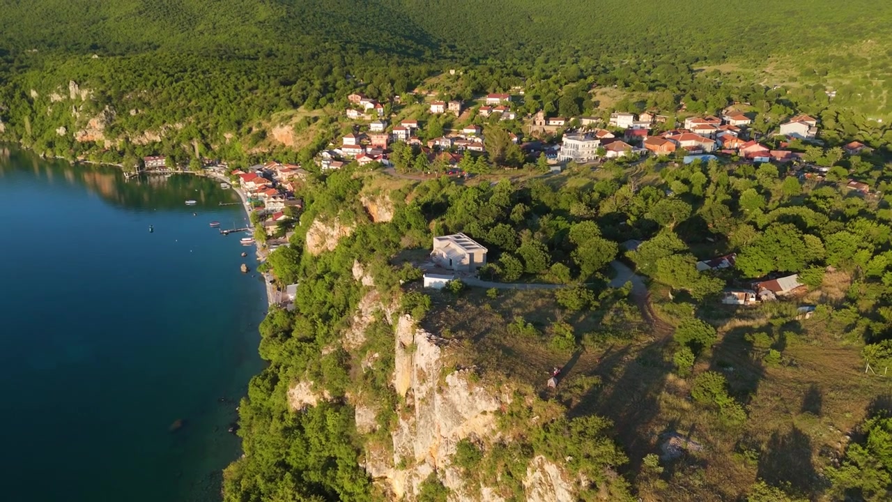 Imagem aérea de drone capturando um assentamento tradicional na encosta, aninhado nas falésias verdes íngremes com vista para o Lago Ohrid. Bela paisagem cultural.