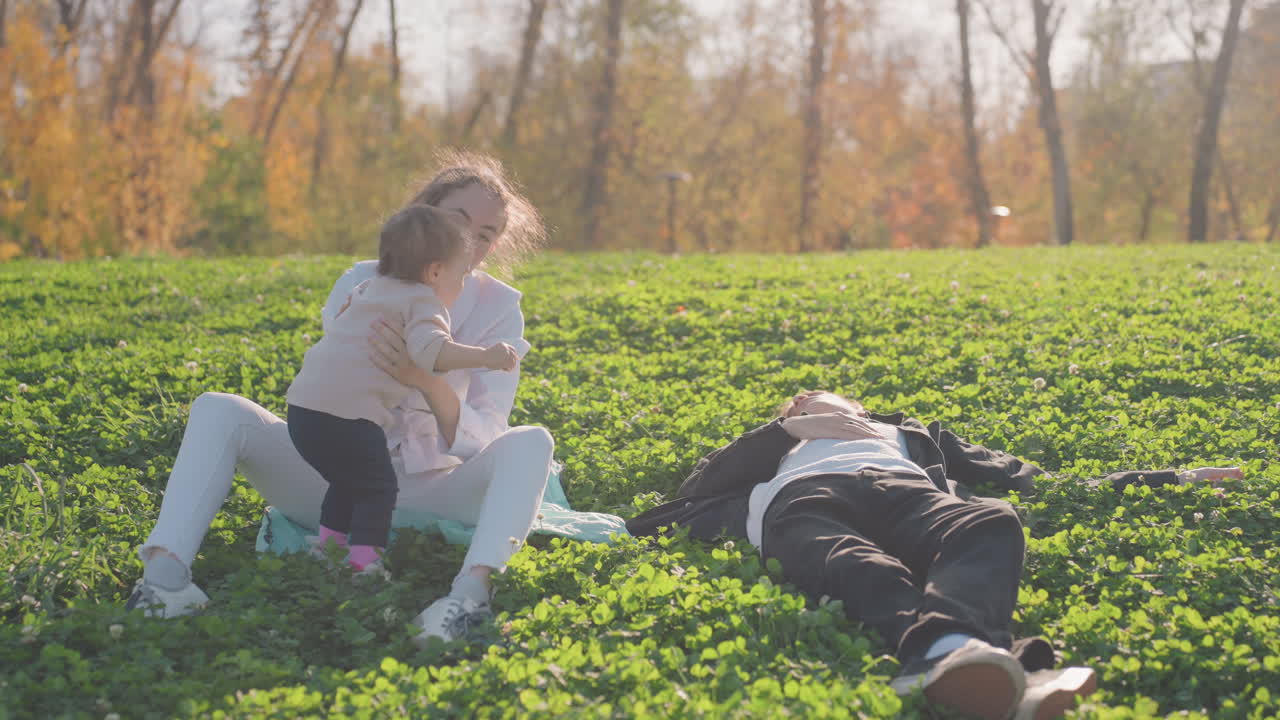 Hvid mor og barn krammer på græs, ægtepar med picnic i baggrunden, hvor faren hviler. Ømt knus og glædeligt kys, gyldent sollys, en intim familiestund, der fejrer omsorg og tillid.