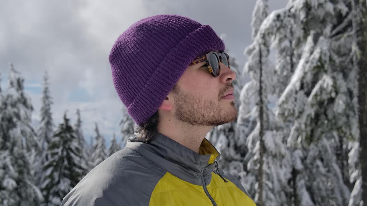 Giovane uomo con berretto e occhiali da sole che guarda gli alberi coperti di neve nella foresta.