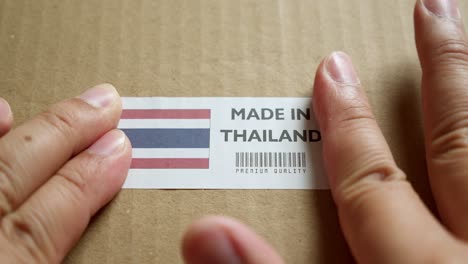 Hände,-Die-Das-Flaggenetikett-„Made-In-Thailand“-Auf-Einem-Versandkarton-Mit-Einem-Barcode-In-Premiumqualität-Des-Produkts-Anbringen