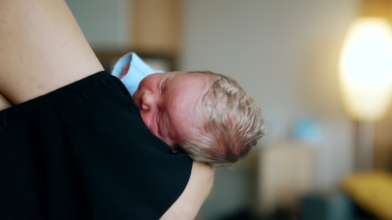 Blond head of a tiny sleeping newborn on mom's arm. Unrecognized mother waving her baby. Close up. Blurred backdrop.