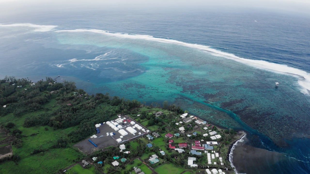 Ripresa aerea con drone del villaggio di Teahupoo, Tahiti, con case costiere accanto al passaggio della barriera corallina dove l'acqua bianca delimita il bordo dell'oceano aperto.