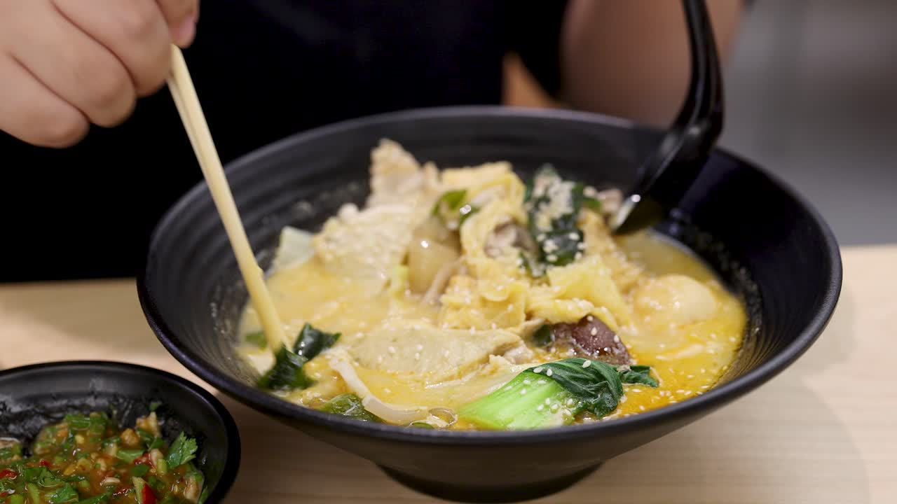 Person enjoys spicy noodle soup with chicken, bok choy, mushrooms, and dipping sauce, close-up view