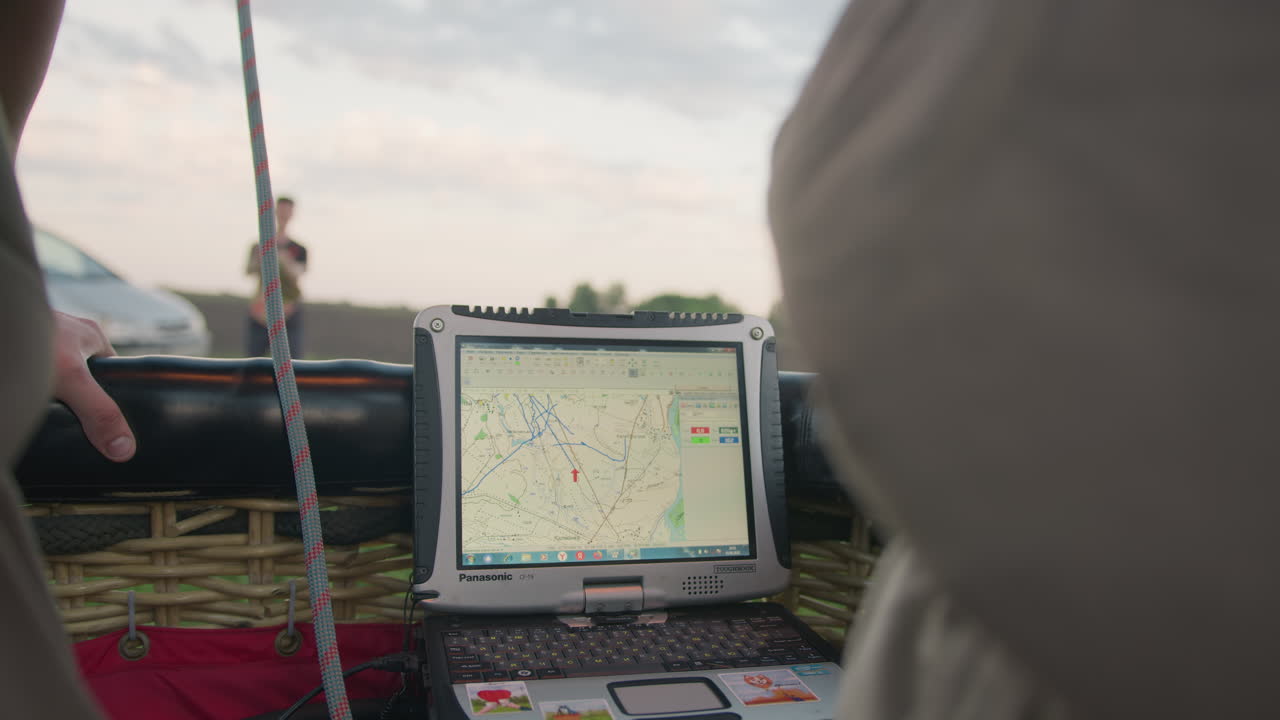 Pilot viewing flight map on rugged laptop mounted in balloon basket, route lines and waypoints visible on display, braided wicker frame and tether rope framing screen with soft evening sky