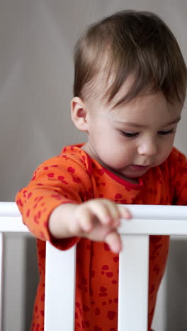 caucasian toddler in orange shirt tries to reach camera with his hand. baby boy standing in the cot drop a blue pacifier and looks how it falls. vertical video