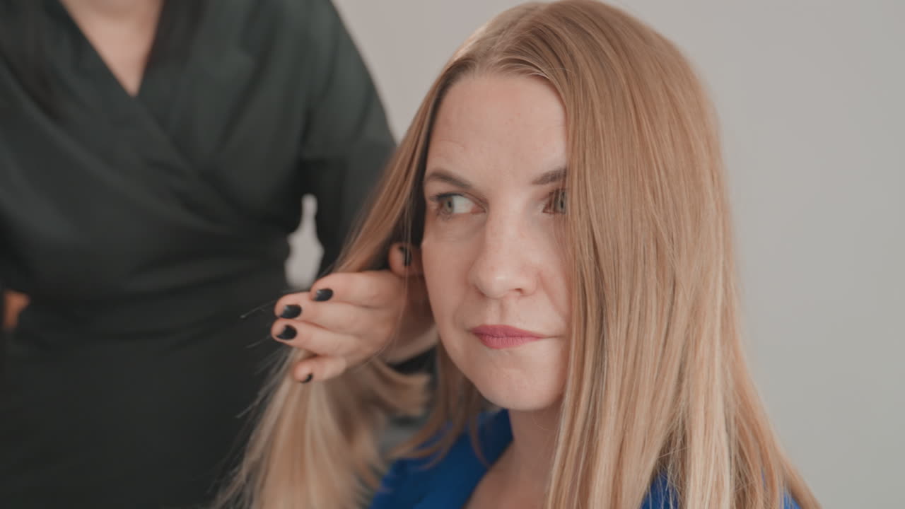 Kaukasische Salon-Stylistin bürstet das Haar, Kundin sitzt ruhig im blauen Umhang, Stylistin glättet verworrene Strähnen mit einer Paddelbürste, Coloristin inspiziert die Spitzen, Model bereitet sich auf das Fotoshooting vor, stille Erwartung