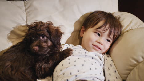 cute chinese kid lies on the couch next to a brown puppy, with the tv