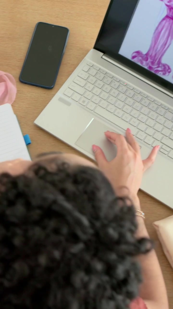 Mujer con blusa color óxido acercándose al escritorio, ajustándose una bufanda rosa, usando una laptop y un cuaderno.
