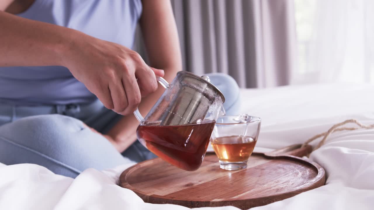 La persona seduta allunga la mano verso la teiera, la solleva e la inclina su un vassoio sul letto per riempire la tazza.