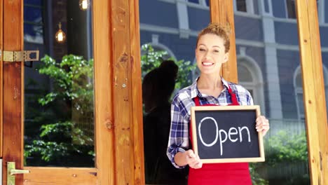 Smiling-waitress-standing-outside-door-with-open-sign-board-in-cafe