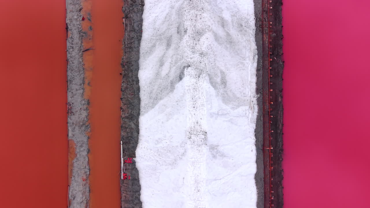 Overhead view of a yellow excavator and white salt pile contrasted against vibrant pink and reddish water ponds. Aerial shot of industrial salt harvesting
