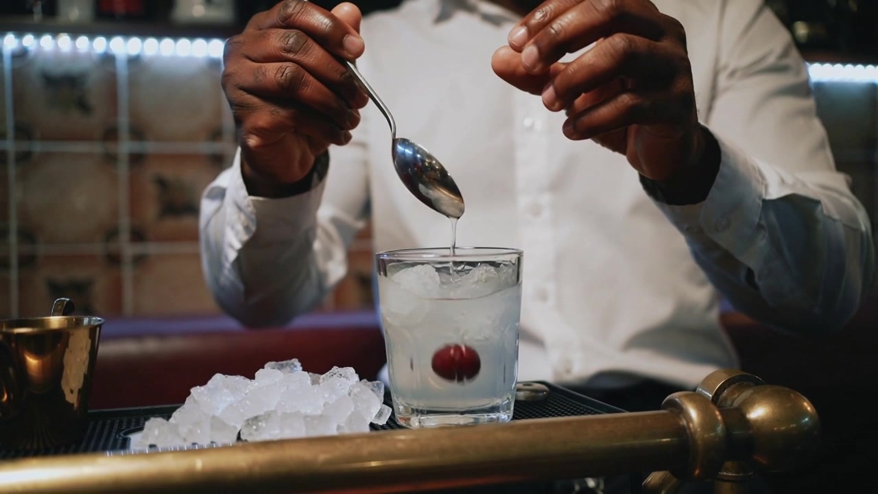 Tilting spoon, white-shirt bartender dropping maraschino cherry into glass at bar