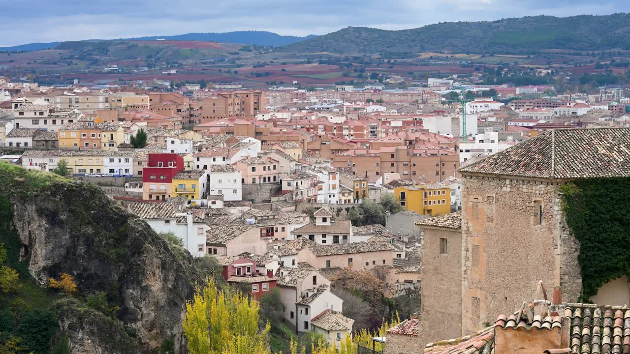 The historic city of Cuenca, Spain, merges with the natural landscape, with its famous buildings perched on cliffs and surrounded by rolling hills on an overcast day.
