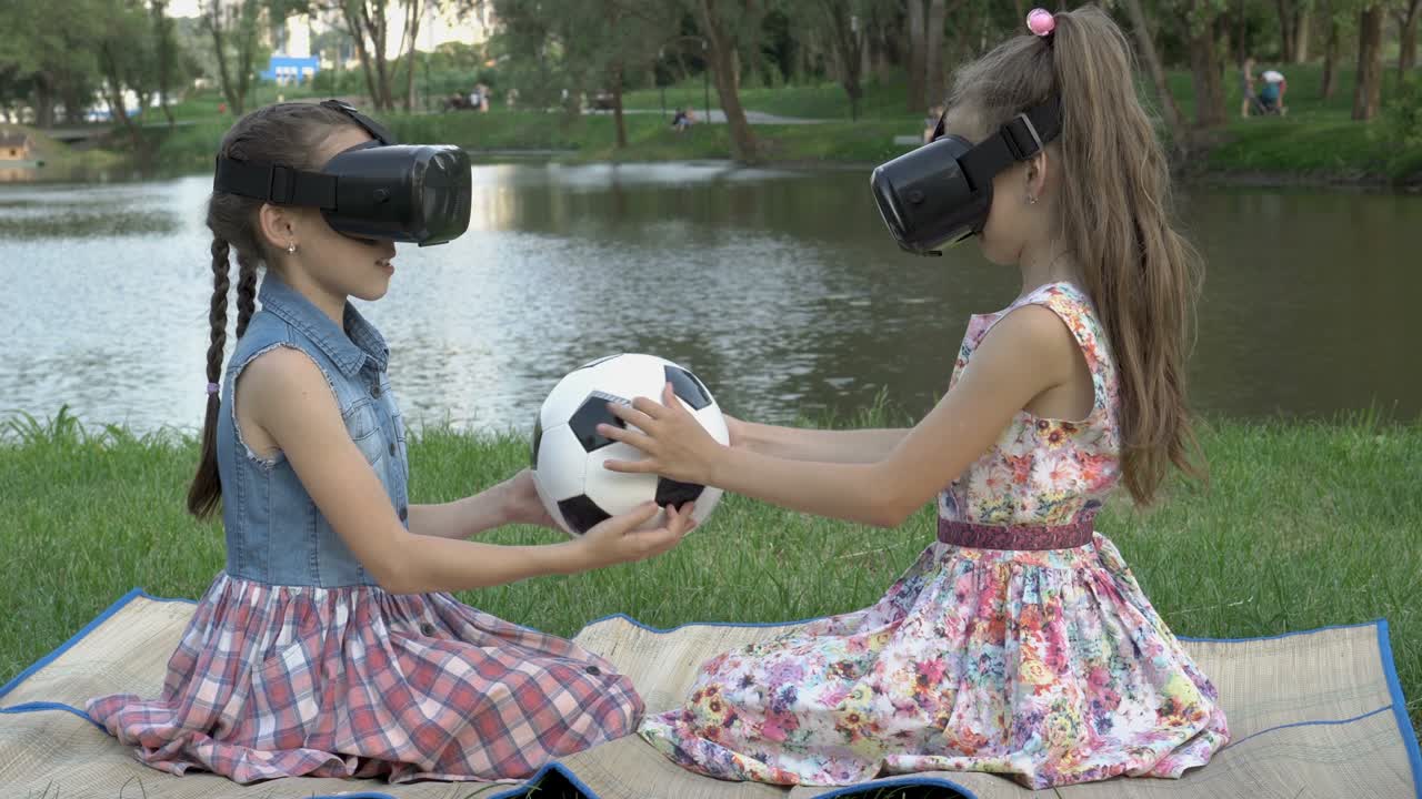 dos chicas jóvenes en gafas de realidad virtual juntas giran y estudian una pelota de fútbol por tacto, sentadas en un parque contra el fondo del agua en un día de verano. el concepto. primer plano. 4k.