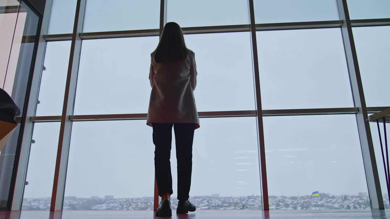 Woman in suit comes up to the big panoramic windows. Thoughtful young lady standing and watching outside. Low angle view.
