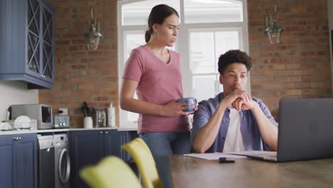 pareja biracial preocupada sentada en la mesa en la cocina usando una computadora portátil