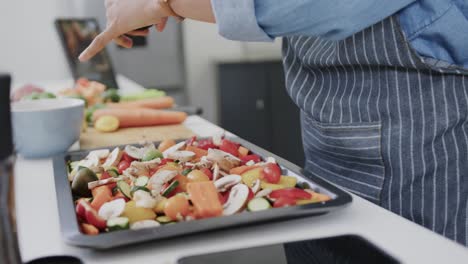 Midsection-of-biracial-woman-seasoning-chopped-vegetables-on-baking-tray-in-kitchen,-slow-motion