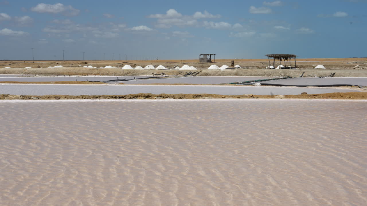Ampla tomada panorâmica das salinas de Manaure em La Guajira, Colômbia, com água ondulante, montes de sal brancos, barracos rústicos e luz do dia clara. Paisagem brilhante e árida, sem pessoas visíveis