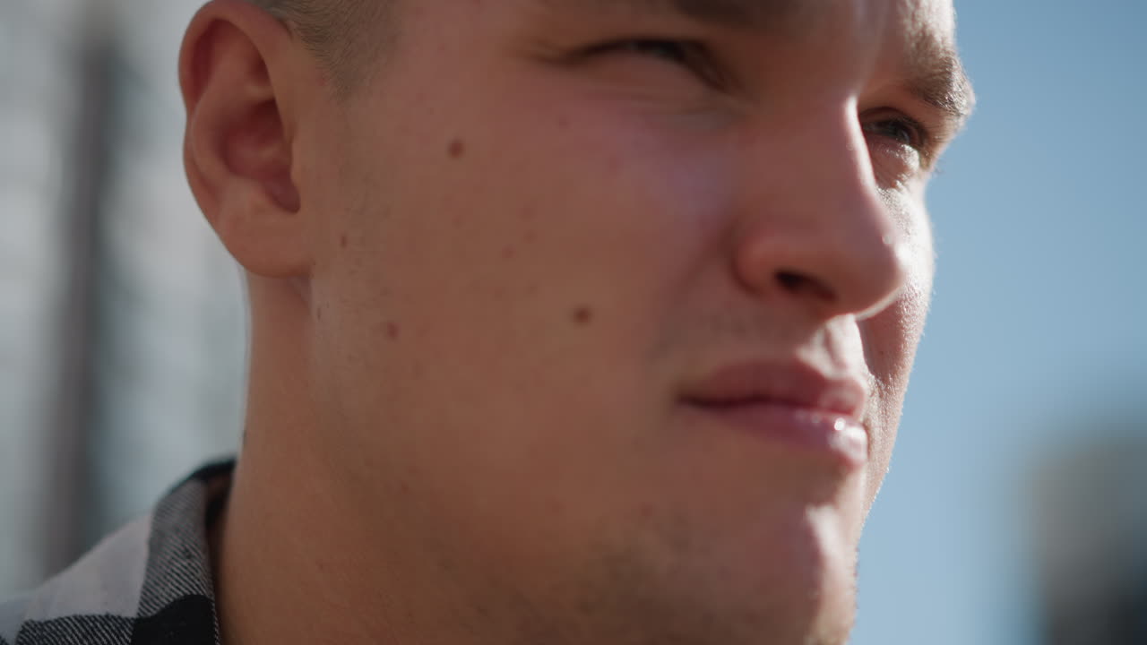 Extreme close up of fair-skinned man with visible facial spots taking something with focused expression as sunlight reflects off face, creating warm contrast in blurred urban background