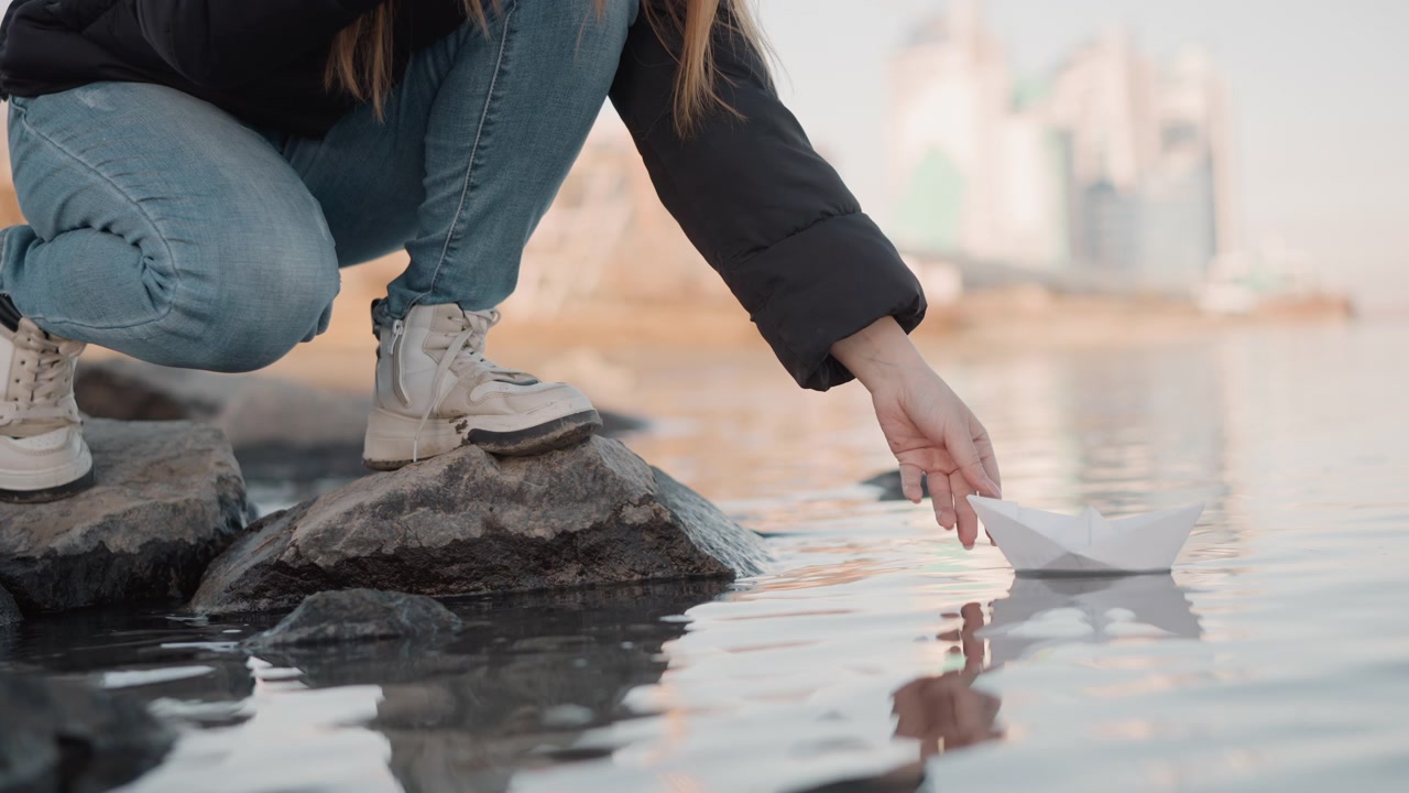 Una giovane donna si accovaccia sulle rocce vicino all'acqua, rilasciando una barchetta di carta in leggere increspature. Indossa abiti casual e scarpe da ginnastica bianche mentre interagisce con l'ambiente circostante in un contesto esterno.