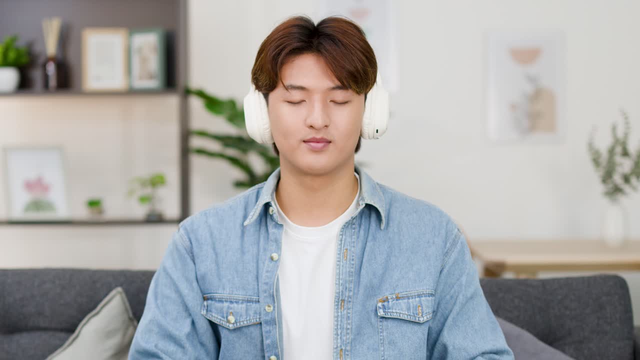 アジア人男性が白いヘッドフォンを着け、ソファで目を閉じて瞑想しています。