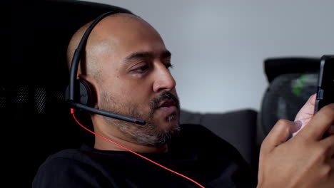 un hombre con auriculares se sienta cómodamente en una silla de ruedas, tocando la pantalla de su teléfono móvil, ilustrando la idea de ocio y entretenimiento en el hogar