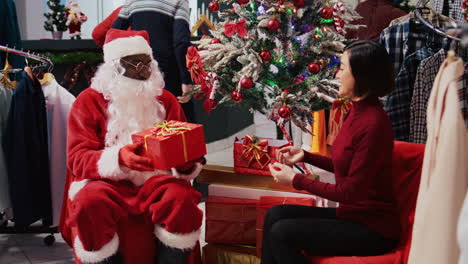 Afroamerikanischer-Manager,-Der-Als-Weihnachtsmann-Fungiert-Und-Neben-Dem-Weihnachtsbaum-Mit-Einer-Frau-In-Einem-Festlich-Geschmückten-Bekleidungsgeschäft-Sitzt.-Leiter-Einer-Modeboutique-Bietet-Einem-Dankbaren-Kunden-Ein-Geschenk-An