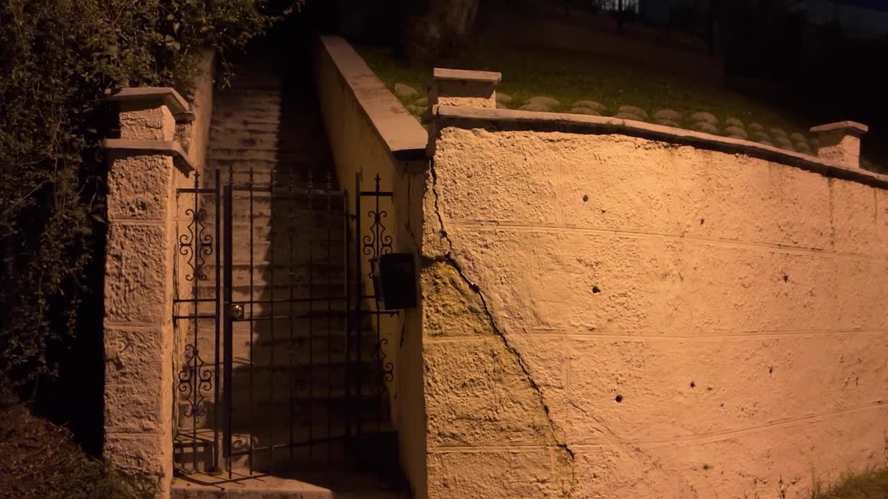 Una toma en primera persona caminando junto a una escalera de piedra con verja y una pared texturizada de noche. Ideal para documentales, suspense urbano o narración de temas sociales.