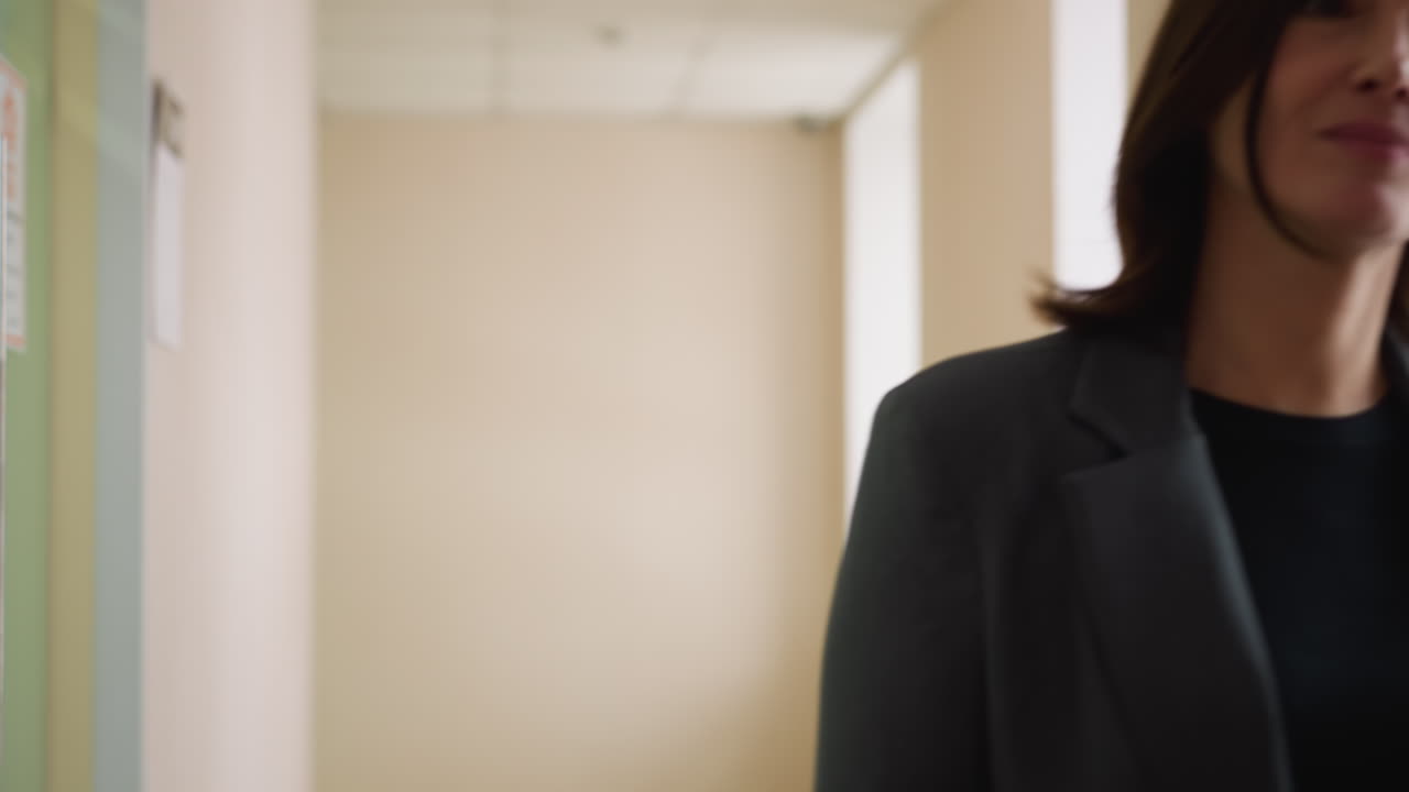Confident businesswoman in gray suit walks toward camera in bright office hallway with composed expression, portraying determination, authority, and calm focus in corporate professional environment