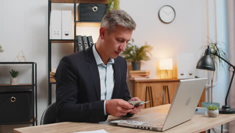caucasian young businessman in suit working on laptop pc counting money cash at home office desk