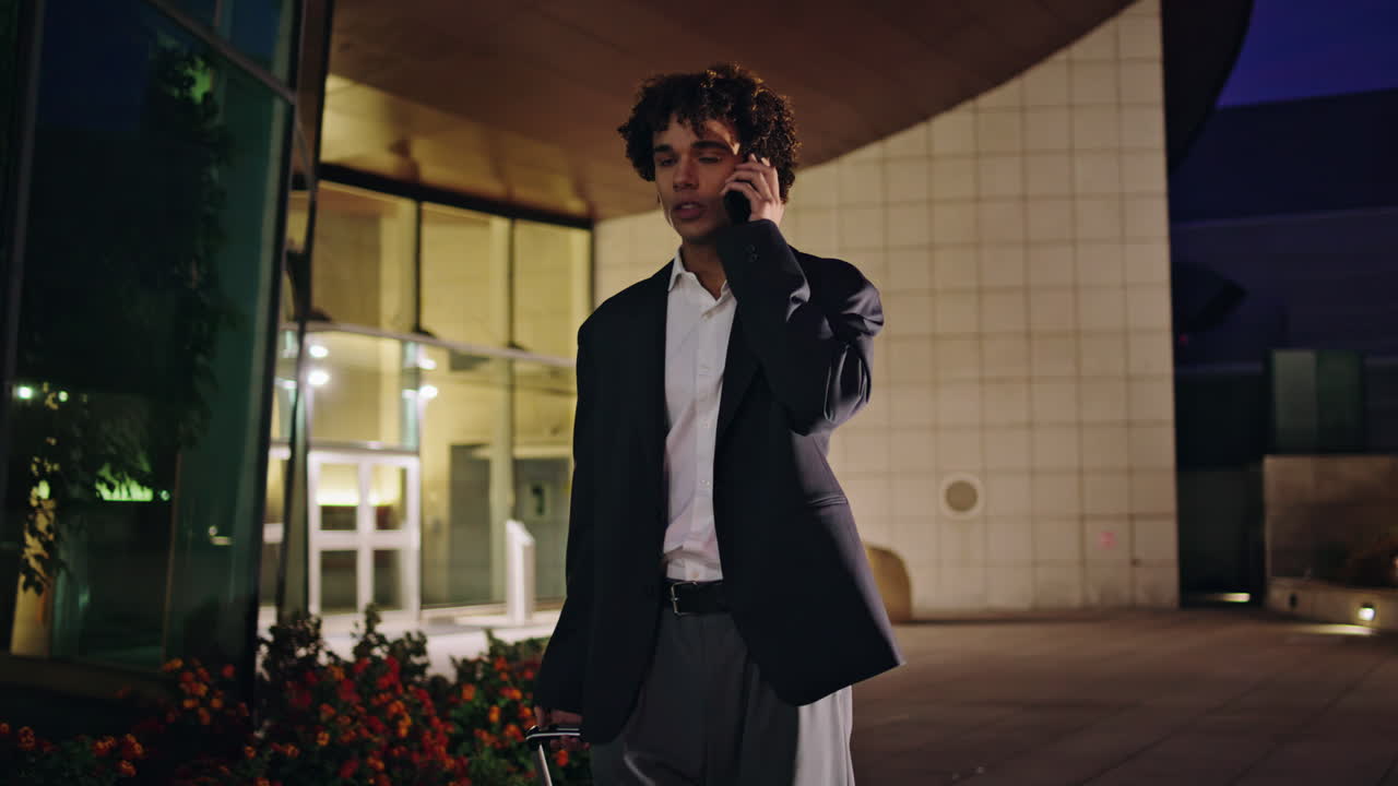 밤 거리에서 짐을 들고 걸어가는 직원. 공식적인 남자가 모바일로 이야기하고 있습니다.
